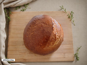 Round loaf of bread on a wooden board with sprigs of greenery and a beige cloth. Text "aikitchen.blog" in the bottom left corner.
