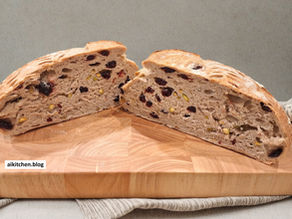 Sliced loaf with cranberries and nuts on a wooden board, gray background. Text on board: aikitchen.blog. Neutral tones.