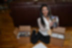 Woman in white blouse and black pants sits on wooden floor, smiling and holding flyers. Surrounded by open boxes, with her print order from wcb5.com