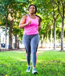 A Black woman with a pink tank, pink ear buds, and grey yoga pants walking briskly through a tree lined park