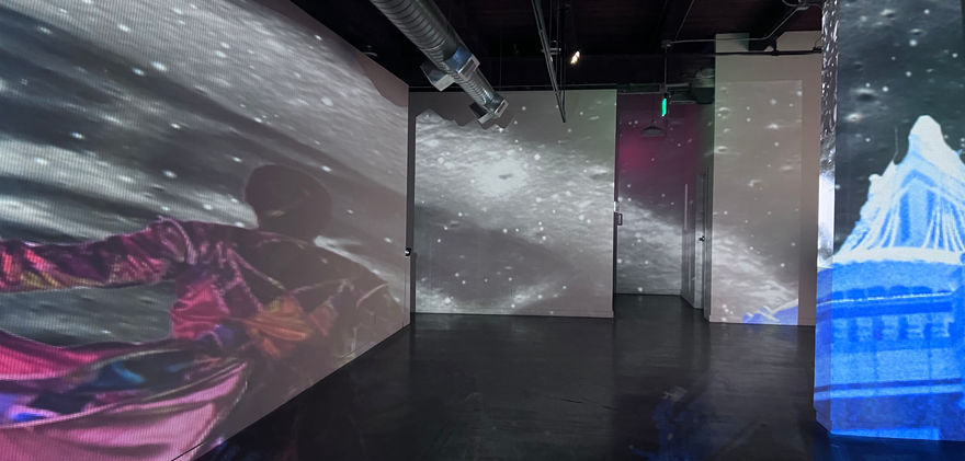 Empty art exhibition shows a woman in a fuchsia dress projected on the left wall. Space projections overlap. Building projected on right wall.