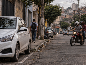 Carro branco estacionado na rua em São Paulo com moto com dois homens passando ao fundo, ilustrando o risco de furto e a importância do rastreamento veicular.