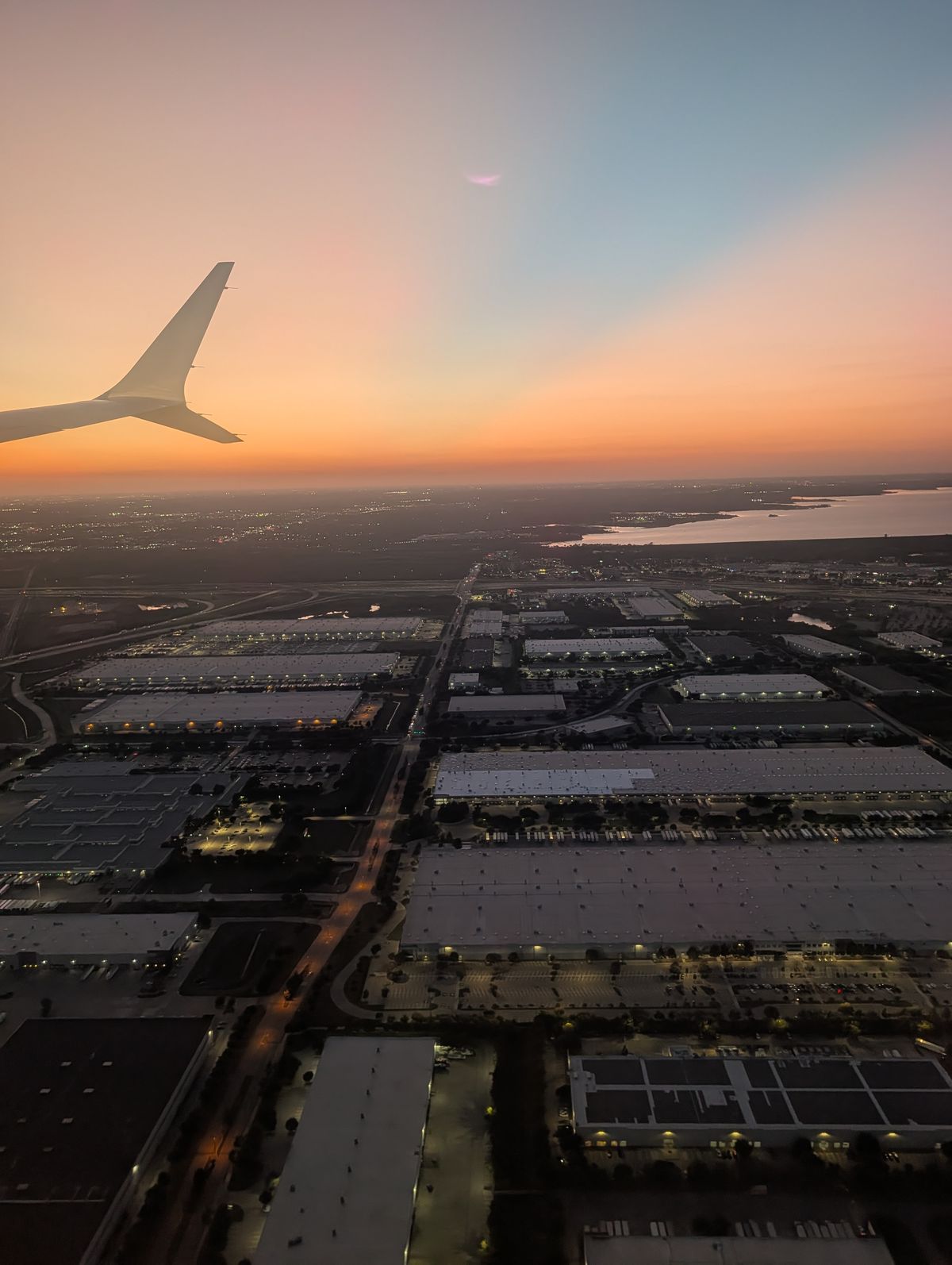 A sunset view from the airplane window, with the wing above buildings, roads and water below.