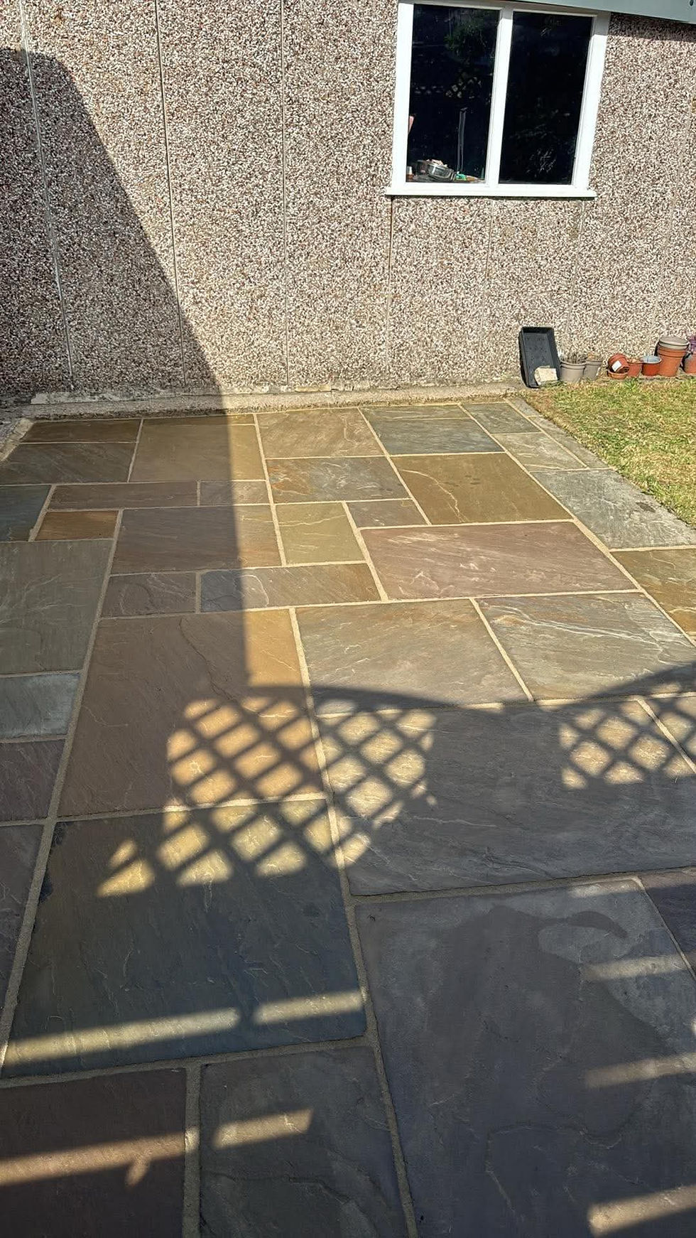 Landscape photo of a clean stone patio in sunlight next to a pebble-dash wall and small lawn, wide landscape orientation, suitable as an example of exterior cleaning results.