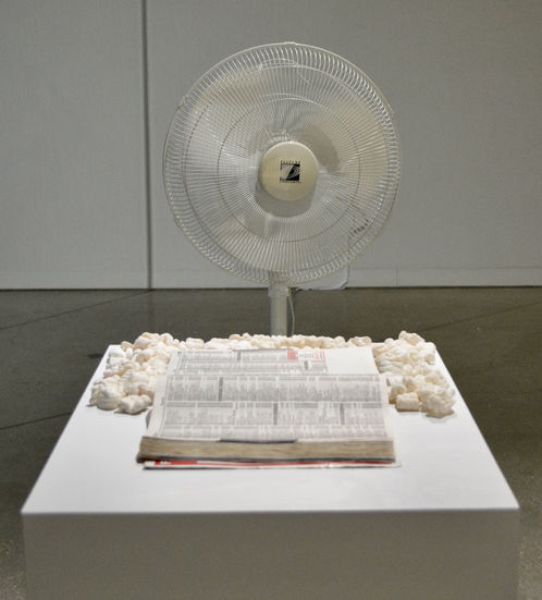 The image shows the head of the fan in the back and the phone book in the front on a pedestal. The phone book has packing peanuts around the half of the book that is nearest the fan.