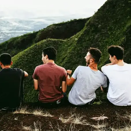 Four male friends laughing and sitting on a rocky mountain ledge during a scenic hike, capturing the importance of friendship, nature, and open conversations in supporting men’s mental health.