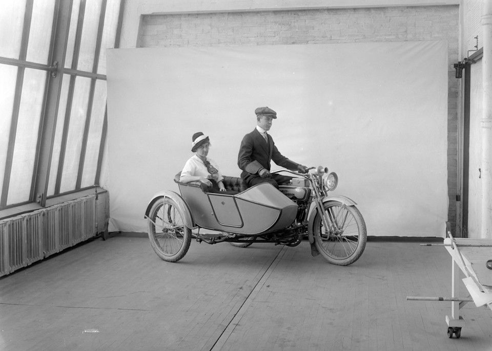 A man sitting on a motorcycle with a woman in the sidecar being photographed in the photo studio. The Motor Company used the medium of photography extensively as a sales tool to promote the benefits of the motorcycle in work and recreation.