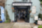 This is the shopfront of the secret bookshelf. The entryway is paved, into a courtyard, before the glass fronted shop entrance. Above the door is a white sign with black writing stating the secret bookshelf. The building is a blue/green colour.
