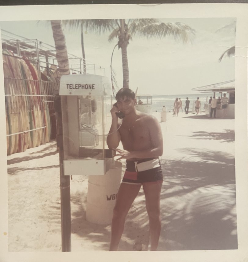 A young man on a payphone at the beach beside a surfboard rack, about 1965