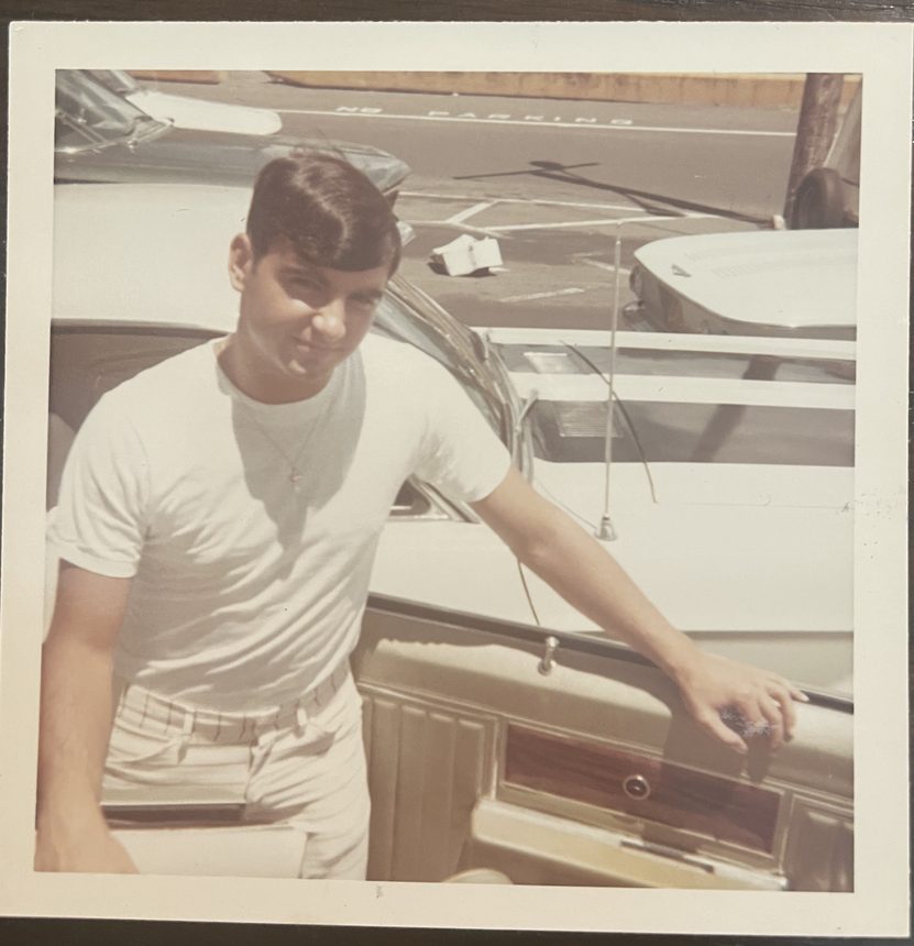 A man leaning on a car fender beneath a no parking sign