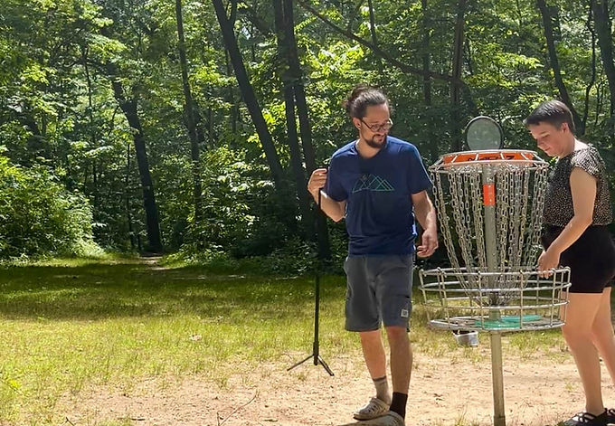 Two people play disc golf at the disc golf basket at the Madeline Bertrand county park.