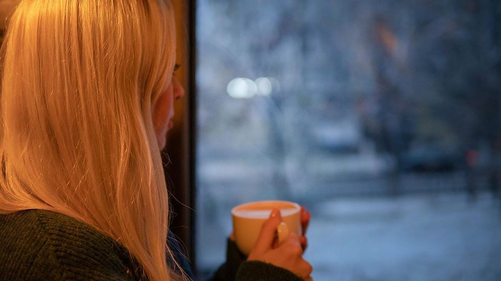 A woman with blonde hair relaxes indoors with a cup of coffee, looking out at the winter scene.