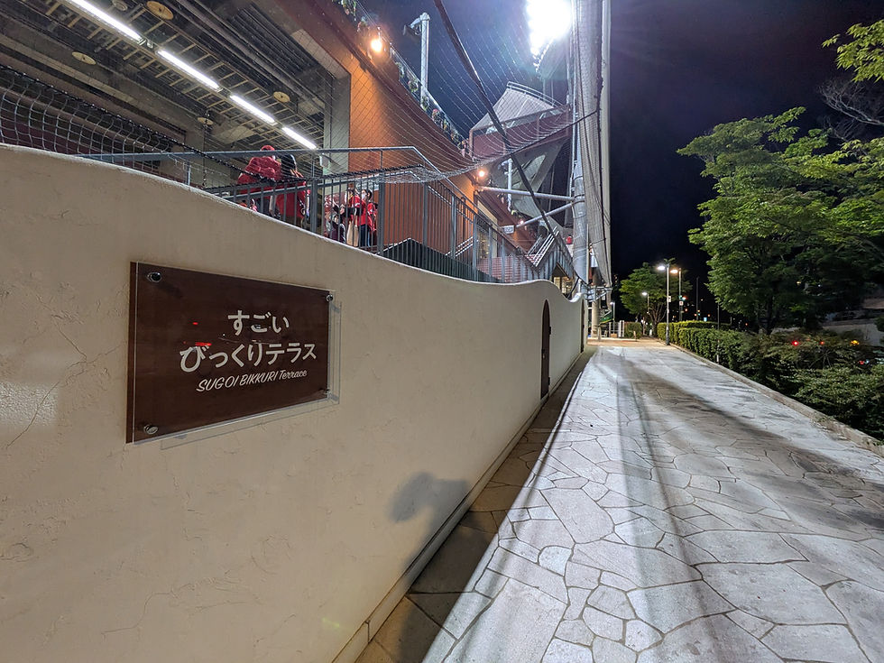 夜のスタジアム外観。白い壁に「すごいびっくりテラス」の看板。数人がテラスで集合。緑の木々が並ぶ道が続く。