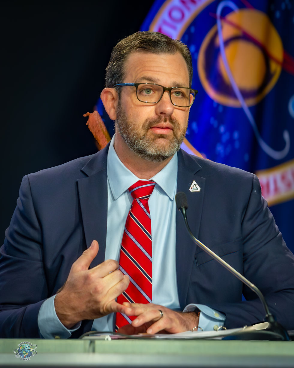 Man in suit and glasses speaks at a podium with microphone, red striped tie, Blue Marble Project logo, blue space-themed backdrop.