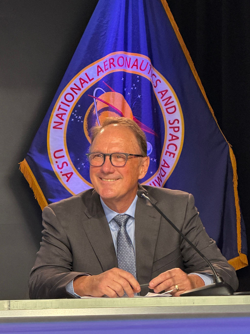 Smiling man in a suit at a podium before a National Aeronautics and Space Administration flag during a formal event
