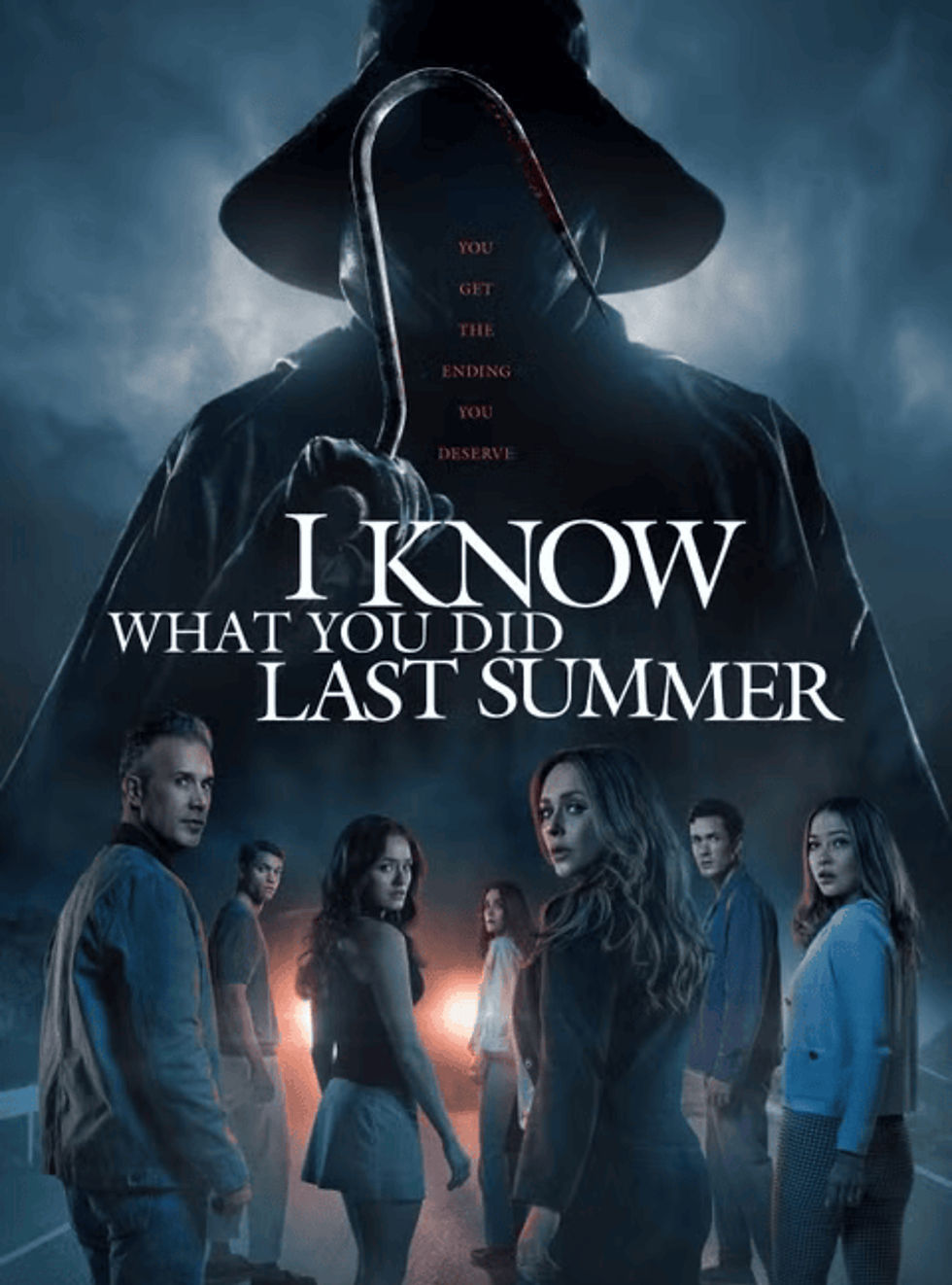 Promotional poster for I Know What You Did Last Summer reboot, featuring a dark, ominous figure holding a hook looming over a group of six young adults standing on a road at night. The tagline reads, “You get the ending you deserve.”