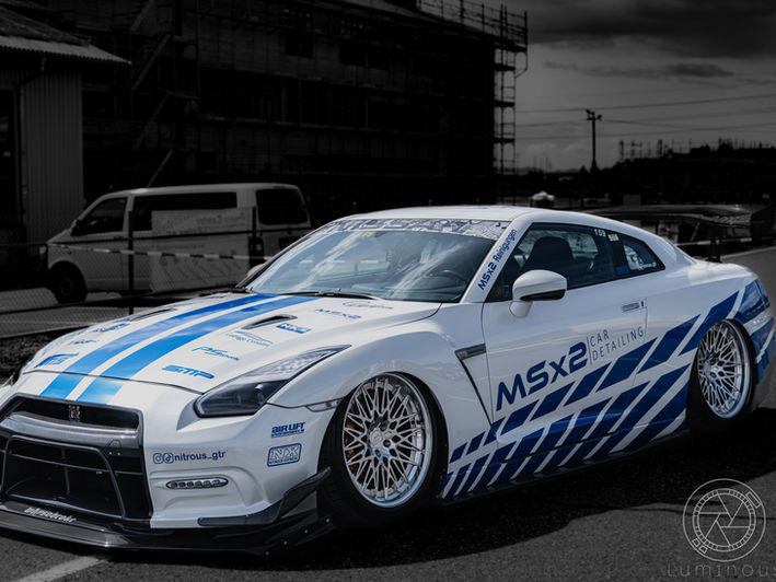 Das Bild zeigt einen getunten Nissan GT-R in einem auffälligen Design, der auf einem Parkplatz steht. Der Sportwagen ist weiß mit blauen Rennstreifen und mehreren Sponsorenaufklebern, darunter "MSX2 Car Detailing" und "Nitrous GTR". Der Wagen hat große, silberne Felgen und einen aggressiven Frontsplitter. Im Hintergrund sind unscharf ein weißer Van und ein Gebäude zu sehen. Das Foto hat einen dramatischen Effekt, bei dem der Wagen farbig und der Hintergrund in Schwarz-Weiß gehalten ist.