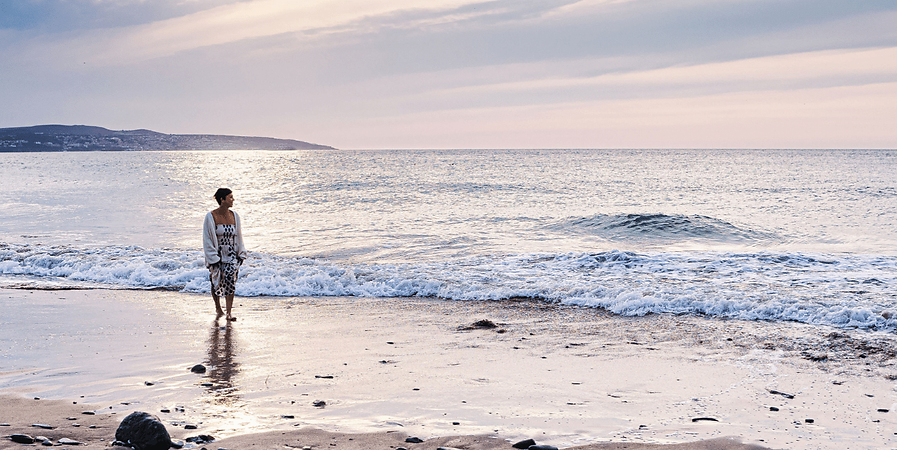 Wide shot of Helen paddling in the shallow sea on a beach with the sunshine reflected in the sea.
