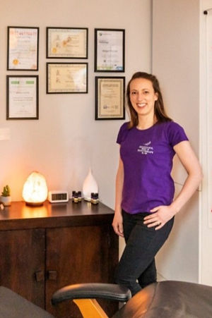 Abi Fenton wears a purple t-shit and black trousers as she stands and smiles in front of her salt lamps and essential oils. The big comfy chair where she offers treatments is also in view