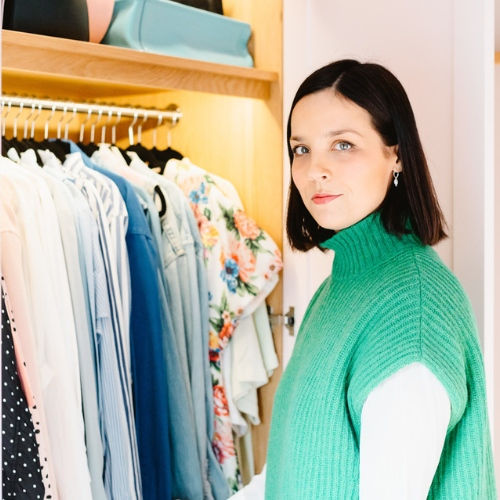 A person in a green sweater stands beside an open wardrobe filled with colorful clothes, looking towards the camera with a neutral expression.