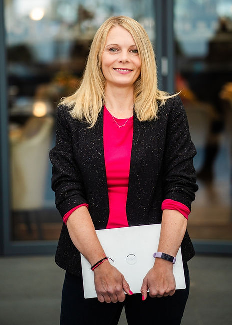 Ladybird SEO owner Jo Jenkins (aka 'SEO Jo') standing up clutching her silver laptop with both hands down in front of her. She has long blonge hair and is facing forward smiling at the camera wearing a black jacket wi the sleeves rolled up with a bright pink top underneath.