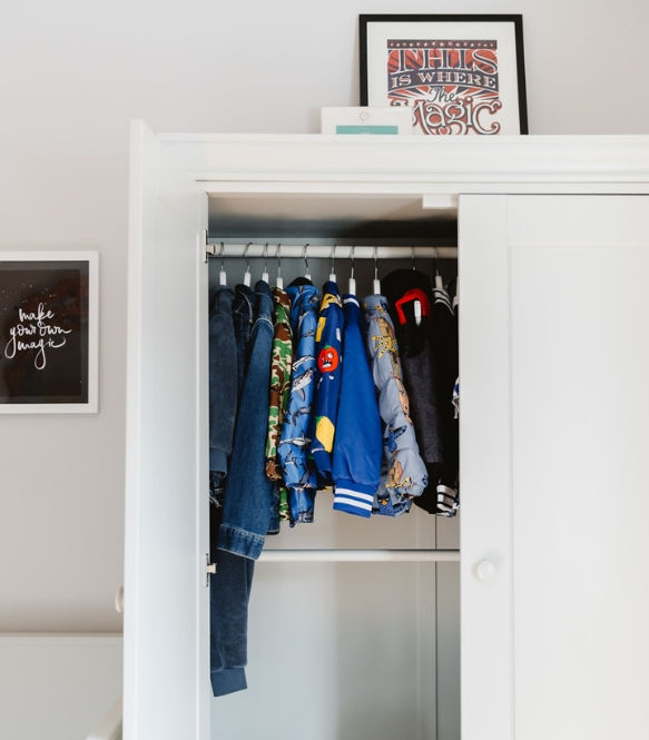 Open wardrobe with colorful children's jackets on hangers. Framed text art on top says "this is where the magic," wall art reads "make your own magic."