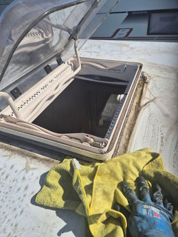 A gloved hand holds a yellow cloth with soapy water as the roof window of a caravan is open and the cloth cleans around it.