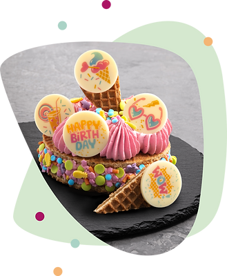 Runde Torte mit rosa Cremehaube, dekoriert mit bunten Zuckerstreuseln und fünf weißen Schokoladentäfelchen mit farbigen Geburtstagsmotiven, arrangiert auf schwarzem Schieferuntergrund.