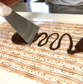 Transferfolie mit braun-goldenem Streifenmuster wird mit flüssiger Schokolade bestrichen.