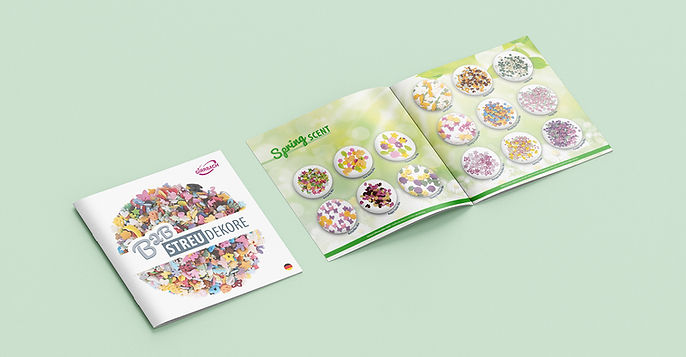 Auf einem hellgrünen Hintergrund liegt links ein geschlossener Produktkatalog mit dem Titel „B2B Streudekore“ von Girrbach. Das Cover zeigt eine große Vielfalt bunter Zuckerstreusel in unterschiedlichen Formen wie Sterne, Herzen, Tiere und mehr. Rechts daneben ist der geöffnete Katalog zu sehen, mit der Überschrift „Spring Scent“. Auf den Doppelseiten sind dekorativ angerichtete Streudekor-Mischungen in runden Präsentiertellern abgebildet, benannt mit frühlingshaften Namen wie „Happy Easter Mix“, „Flower Mix“, „Spring 1“ usw. Das Layout ist hell, freundlich und saisonal gestaltet.