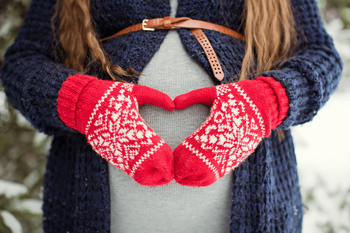 Pregnant woman wearing red mittens, forming a heart shape with her hands over her baby bump, celebrating the holiday season with love and warmth.