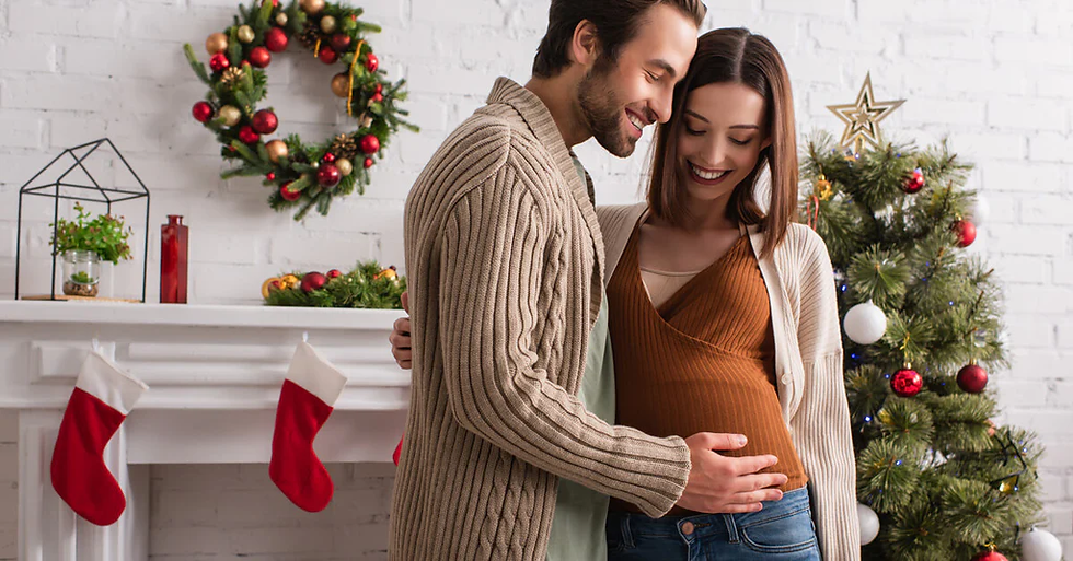 A happy couple celebrating the holidays, with the man gently placing his hands on the pregnant woman's belly, surrounded by a Christmas tree and festive decorations in the background, perfect for a joyful holiday pregnancy moment.