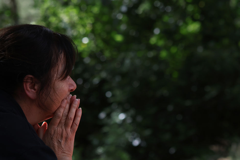 Femme de profil, mains jointes près de la bouche, sur fond vert flou, air pensif. Le retranchement au fil du chemin : du mourant à l'endeuillé.