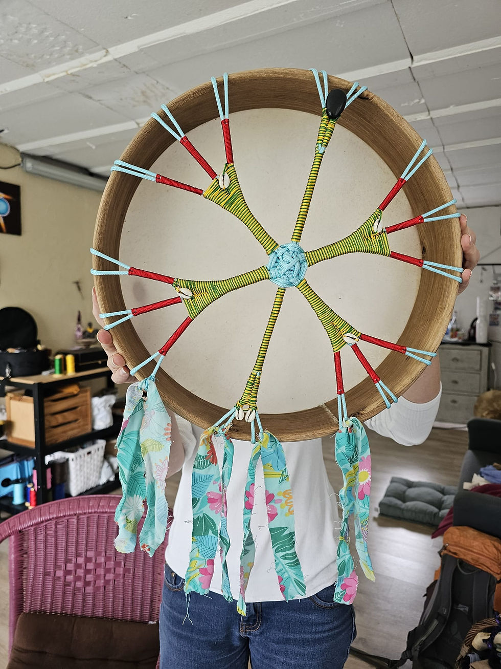 En intérieur, une personne, semblant être une femme tient son tambour chamanique devant elle, cachant le haut de sa poitrine et sa tête. Le tambour est au centre de la photo, cordage visible. Le coeur du tambour fait en corde synthétique est de couleur bleu turquoise clair. 6 branches partent du coeur, en symétrie deux se font face de chaque côté du coeur, elles son fines et tressées de deux couleurs interverties : jaune et vert. Au bout de chaque branche, deux petits coquillages blancs. Les quatre autres branches ont la même structure, elles sont plus larges, les couleurs sont les mêmes et s'intervertissent également. Chacune des quatre branches se séparent au bout en trois, le tissage est alors rouge. Au départ de ces trois petites branches très fines, rouges, des coquillages semblables à ceux placés sur les deux premières branches. En bas du tambours, des bandes de tissus accrochés aux branches tombent à l'extérieur et flottent dans le vide. Ils sont de couleurs bleu turquoise clair, portent des fleurs roses et jaunes ainsi que des feuilles vert foncé. La personne portant le tambour est en jean et porte un tee-shirt manches longues blancs. Le plafond de la salle est en polystyrène, les murs crépis sont couleur sable. Près de sa jambe droite une chaise en osier, rose. A sa gauche, un coussin au sol.