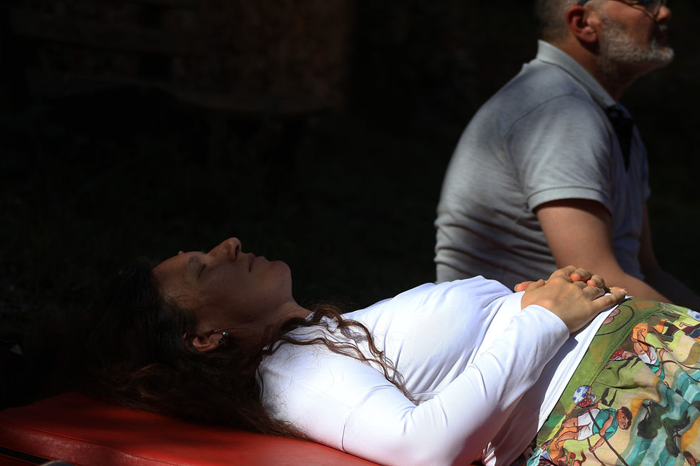 Gros plan de profil d'une femme défunte reposant paisiblement sur un lit de veillée funèbre, les yeux fermés et les mains jointes sur son abdomen. Elle est vêtue d'un haut blanc et d'une jupe colorée. En arrière-plan, plongé dans l'ombre, un homme barbu est assis en silence, assurant une veillée de prière et de recueillement. La scène est éclairée par une lumière naturelle douce, symbolisant le passage et le deuil. Crédit or-photographie