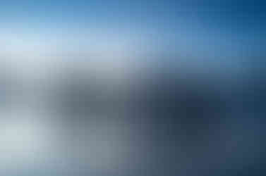 Lever de soleil à la plage de la perle en Guadeloupe. La colométrie de la photo est dans les bleus grisâtres. Photo en format paysage. Le paysage une plage avec une petite vague en formation. A l'horizon, une légère lueur oranger laisse entrevoir l'arrivée du soleil. Bande de nuages gris très foncé dessinant des cheminées, six. Au-dessus le ciel presque blanc tend peu à peu vers un bleu azur. Dans l'eau, dix personnes se donnent la main formant une ligne parallèle à l'horizon et à la vague en formation. L'obscurité de leurs silhouettes ne permet pas de préciser le genre des personnes présentes sur la photo.