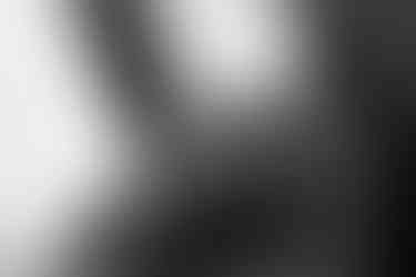 Photo noir et blanc. Zoom sur deux femmes. Fond de la photo est blanche avec un poteau penché vers la gauche, il est foncé. A droite, le bas du visage , on voit les narines et sa bouche fermée. Elle sa main droite sur le front de la femme devant elle. Le haut du visage de la deuxième femme est sous le menton de la première. Leurs visages sont tournés dans la même direction. On voit son regard dans l'ombre. Sa main droite est posée sur celle de l'autre femme sur son front. Elle a les doigts écartés. A son poignet trois bracelets de perles rondes. Deux sont blancs et au milieu le troisième est foncé. Le bracelet clair éloigné de la main porte un coeur ceux au centre, argenté.