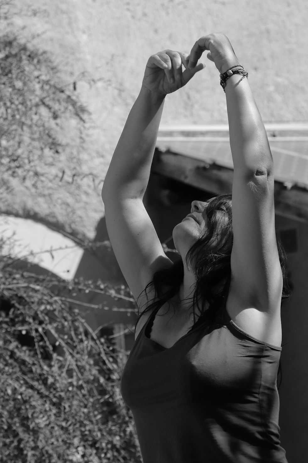 Photo en noir et blanc, en fond les murs d'une vieille bâtisse, avec un arrondi laissant imaginer un dessus de porte, également un arbre style buisson. Une femme qu'on voit à partir du buste, a les bras levés vers le ciel, les mains se touchent formant un ovale au-dessus de sa tête renversée en arrière. Elle porte un tee-shirt à fines bretelles et des bracelets à son poignet gauche. Elle est de face.