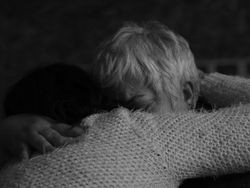 Gros plan en noir et blanc d'une personne âgée aux cheveux blancs, vue de dos, étreignant fermement une personne plus jeune dont le visage est caché. On distingue la main de la personne âgée agrippant le pull en tricot de l'autre personne. L'image capture une étreinte intense et silencieuse, illustrant la douleur invisible du deuil blanc et de la mort sociale, des thèmes abordés par une thanadoula. Crédit or-photographie