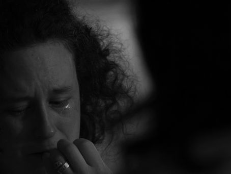 Gros plan en noir et blanc d'une femme aux cheveux bouclés, le visage marqué par l'émotion avec des larmes qui coulent sur ses joues. Elle porte sa main à sa bouche dans un moment de lâcher-prise. Cette image illustre les larmes d'émotion et de relâchement du système nerveux, essentielles pour l'apaisement et le retour à l'équilibre lors d'un deuil ou en fin de vie. Crédit or-photographie