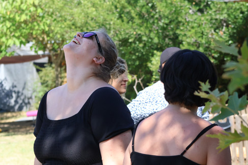 En extérieur, dans un jardin aux arbres verts clairs et au sol d'herbe du même vert. On devine au fond à gauche un bâti gris clair. Au premier plan, deux femmes , une de face l'autre de dos. On devine d'autres personnes derrière elles, sans pouvoir les décrire. La femme de dos, aux cheveux noirs et courts porte un débardeur noir à fines bretelles. La femme de face à sa gauche est plus grande. Elle est blonde, porte des lunettes de soleil noire et une boucle d'oreille dorée à l'oreille gauche. Ses cheveux sont attachés, elle a la tête basculée en arrière et rigole. Sa bouche est ouverte et on voit les dents de sa mâchoire supérieure. Elle porte un tee-shirt noir manches courtes.