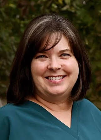 Woman with dark hair and teal scrubs smiles for a professional headshot outside against a green nature background