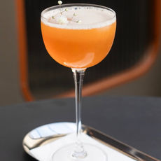 Drink autoral de cor laranja servido em taça, decorado com pequenas flores.