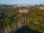 Vista aérea do Parque Tanguá, em Curitiba, mostrando o mirante terracota no topo de um paredão de rocha, de onde desce uma alta cascata em direção a um lago, cercado por vegetação.