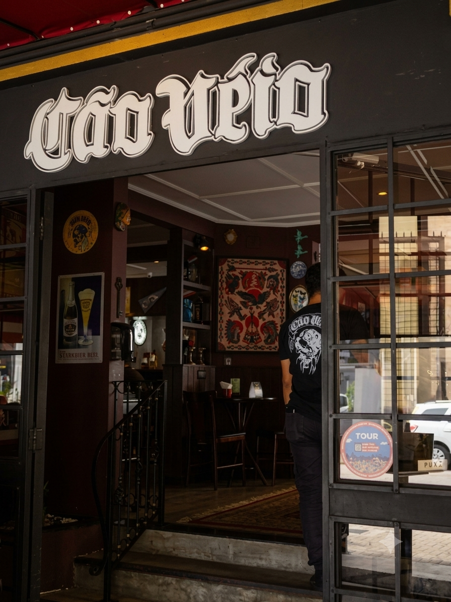 Uma fotografia nítida e bem iluminada mostrando a entrada do restaurante Cão Véio. O letreiro branco e estilizado "Cão Véio" se destaca em uma fachada preta acima das portas de vidro com grade metálica. Através das portas, o interior acolhedor com decoração vintage é visível, incluindo pôsteres de cerveja, obras de arte na parede e mesas e cadeiras de madeira. Uma pessoa de costas usando uma camiseta preta com uma estampa está em pé perto da entrada. Um adesivo "TOUR" com o logotipo do Cão Véio e um adesivo "Puxa" estão no vidro da porta direita. O exterior tem um toldo vermelho e um detalhe de borda amarela.