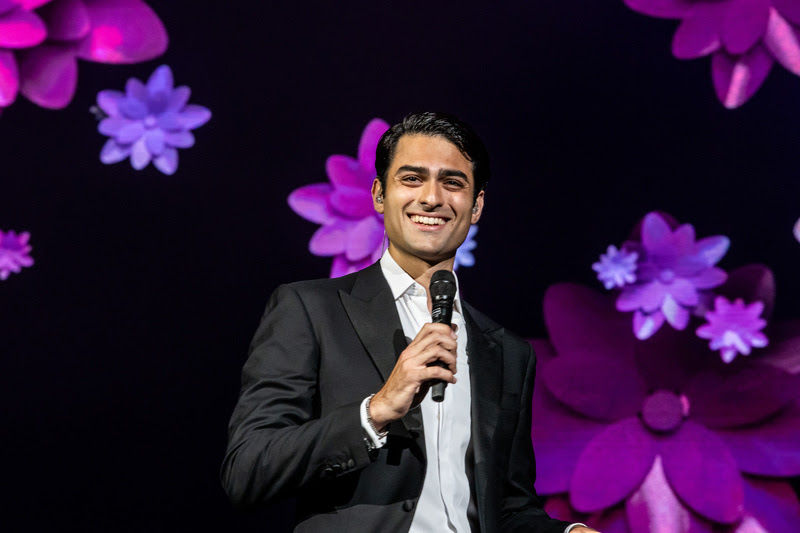Matteo Bocelli se apresenta no palco segurando um microfone diante de um cenário com flores em tons de rosa e roxo.