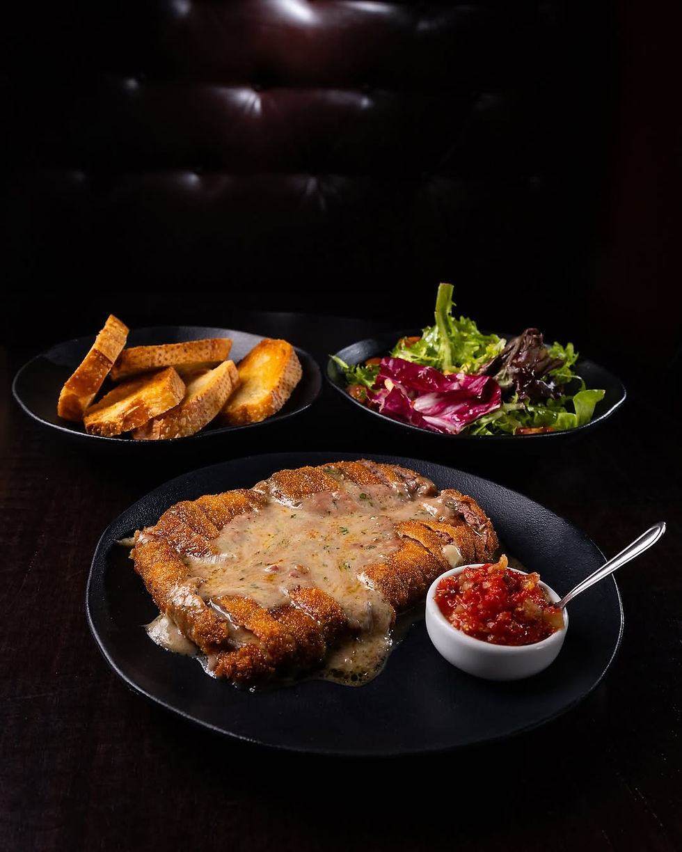 A imagem do arquivo "Cao Veio - Fila Brasileiro (1) (1) (2).jpg" exibe, sob iluminação dramática, uma composição culinária rústica. No centro, sobre um prato preto, está um grande bife à milanesa fatiado, regado com um molho cremoso de cor amarelada. Ao lado, em um pequeno pote branco, há molho de tomate picado e uma colher de metal. No fundo, duas outras tigelas pretas apresentam acompanhamentos: uma com fatias de pão tostado e outra com salada de alface e radicchio. O fundo de parede preta é escuro e acolhedor.