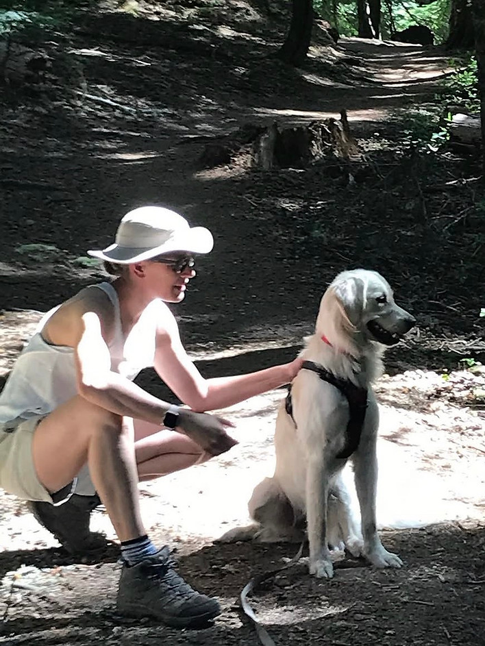 Person kneels on the ground with their hand on the back of a white dog that is sitting. They are on a shady trail in the woods.