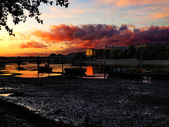 Sunset over calm river Thames with silhouetted boats and a bridge, warm orange clouds reflecting on the water