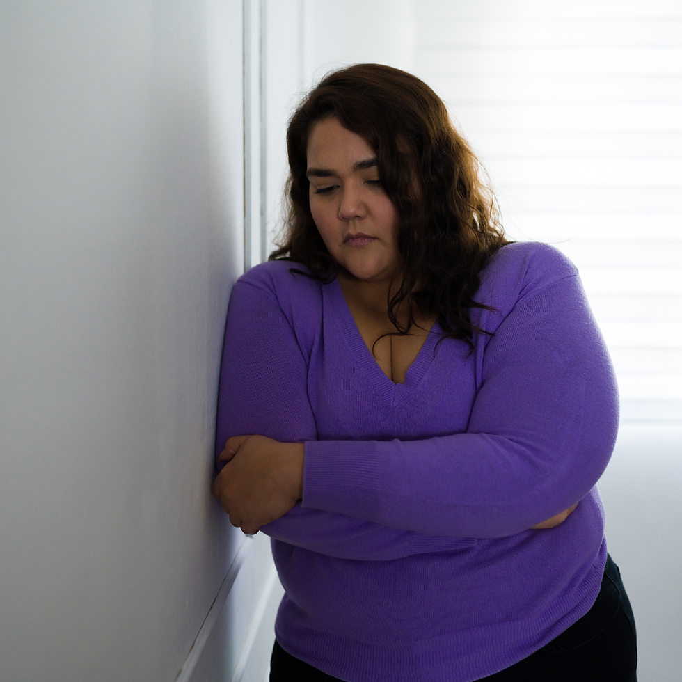 Woman in a purple sweater leans against a white wall with crossed arms, looking pensive. Soft light from a nearby window.